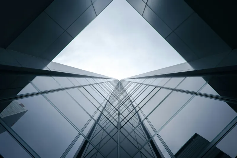Looking straight up through a glass-and-steel atrium roof, its grid of panels converging toward bright daylight