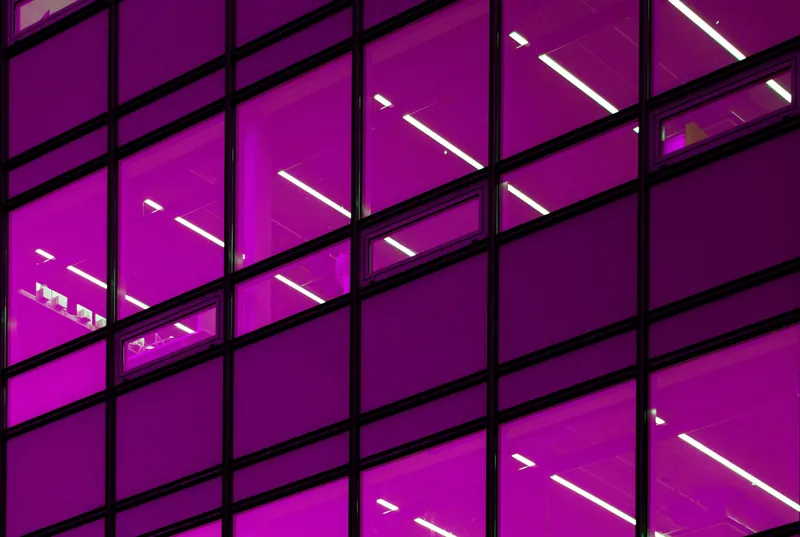 An angled violet glass facade laid out in a repeating grid, lit from within by warm bands of light